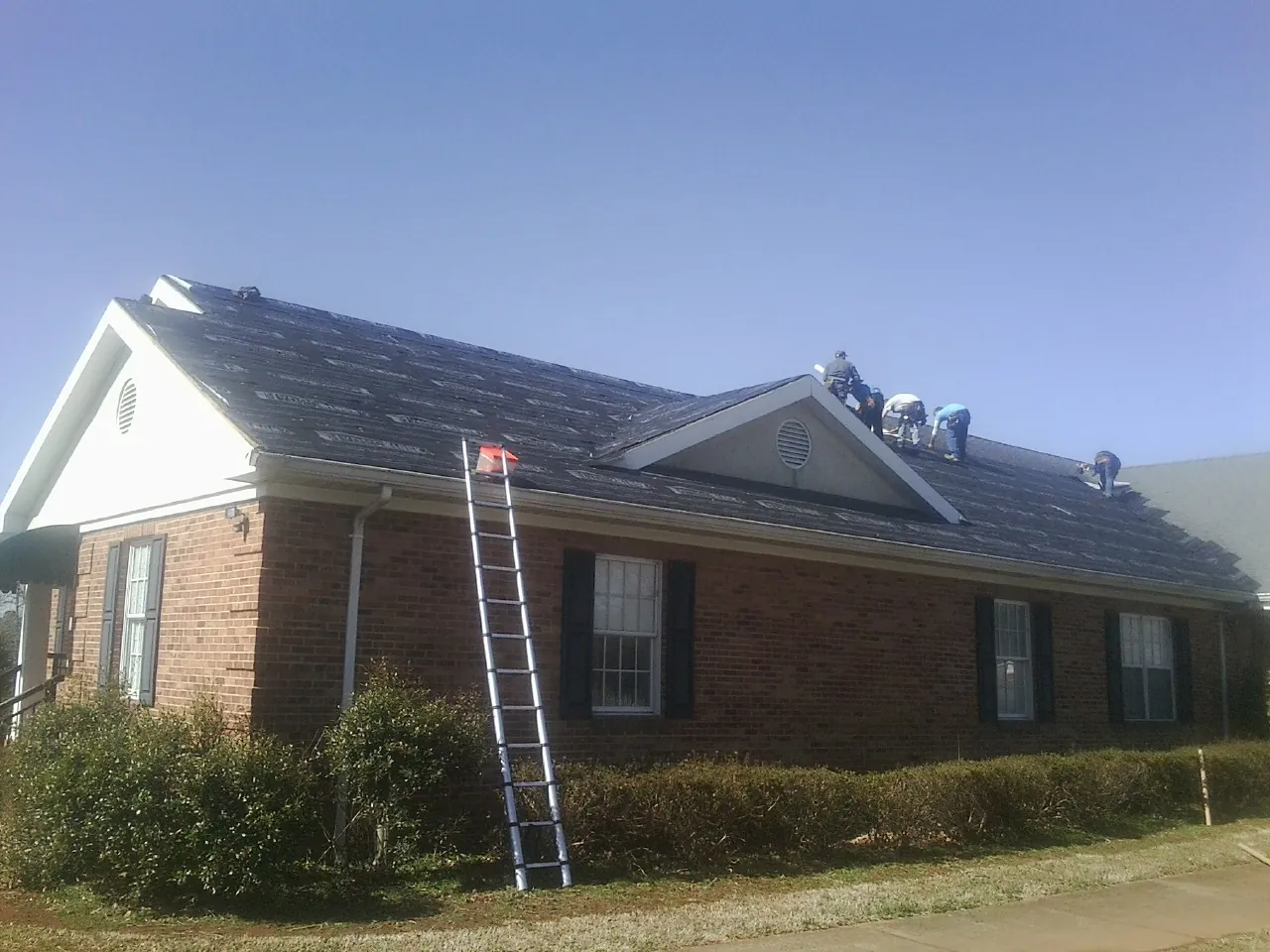 Crew tearing shingles off a brick ranch roof with a ladder set — Emergency Roof Repair work by our crew in Keuka Park