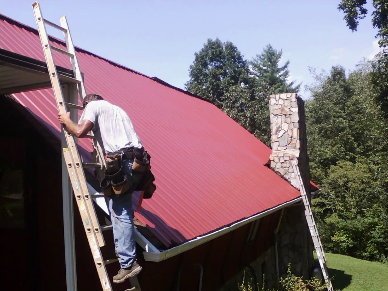 Roofer on a ladder beside a red metal roof and stone chimney — Soffit & Fascia Repair by Roofing Brothers in Keuka Park